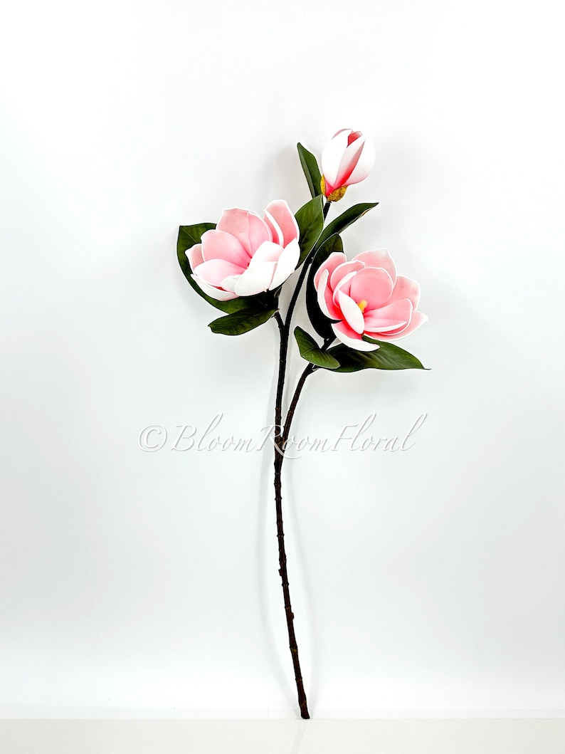 May include: A single stem of artificial pink magnolia flowers with green leaves. The stem is brown and has a natural look.