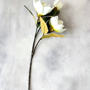K&ouml;nnte beinhalten: Ein k&uuml;nstlicher Magnolienstiel mit drei wei&szlig;en Bl&uuml;ten, gelben Zentren und gr&uuml;nen Bl&auml;ttern. Der Stiel ist braun und strukturiert. Der Text "&copy; Bloom Room Floral" ist sichtbar.