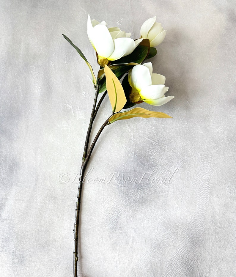 K&ouml;nnte beinhalten: Ein k&uuml;nstlicher Magnolienstiel mit drei wei&szlig;en Bl&uuml;ten, gelben Zentren und gr&uuml;nen Bl&auml;ttern. Der Stiel ist braun und strukturiert. Der Text "&copy; Bloom Room Floral" ist sichtbar.