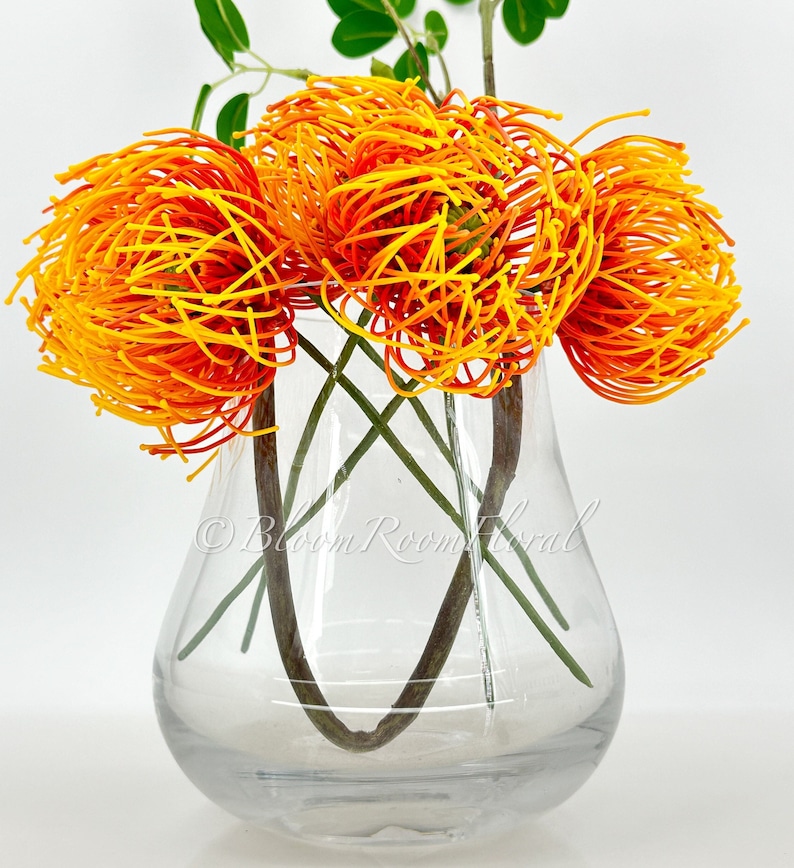 K&ouml;nnte beinhalten: Drei k&uuml;nstliche Pinienzapfenblumen in Orange- und Gelbt&ouml;nen in einer klaren Glasvase. Die Vase ist rund mit einem breiten Boden und einem schmalen Hals.
