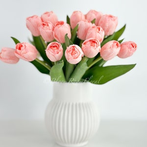 May include: A bouquet of pink tulips in a white ceramic vase with vertical ridges. The vase is sitting on a white surface.