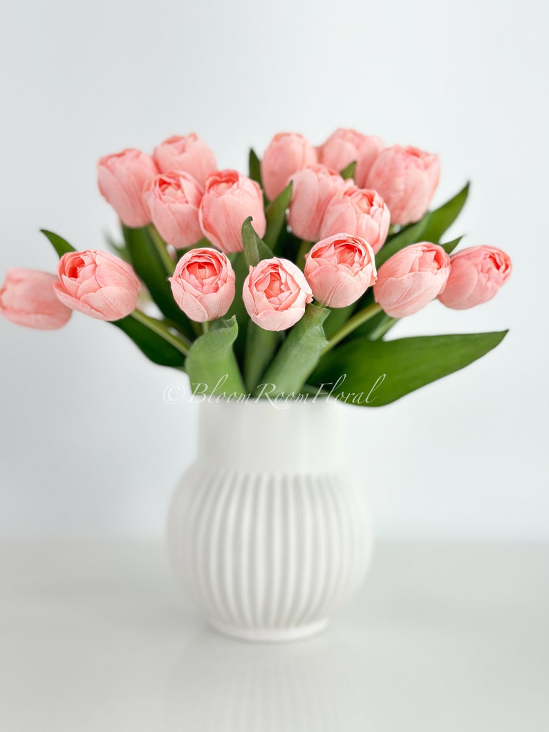 May include: A bouquet of pink tulips in a white ceramic vase with vertical ridges. The vase is sitting on a white surface.