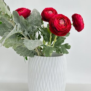 May include: A white, textured vase holds a vibrant arrangement of red ranunculus flowers and silvery-green foliage. The vase has a geometric diamond pattern. The flowers are in full bloom, showcasing their layered petals.