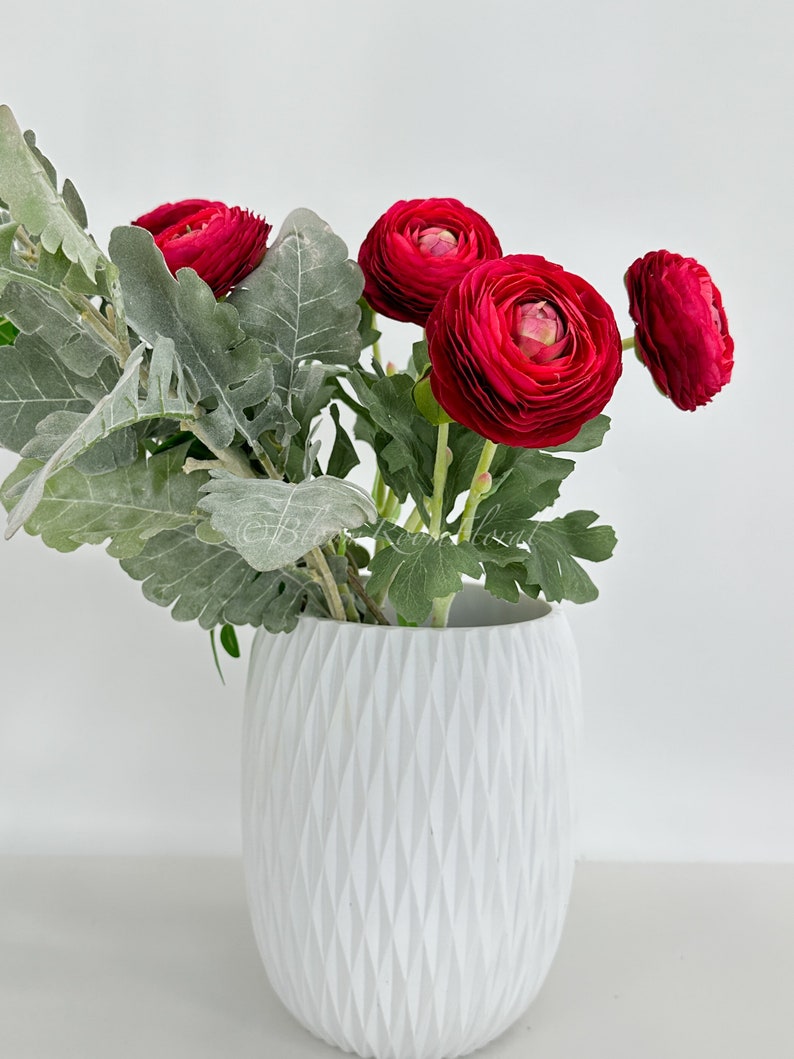 May include: A white, textured vase holds a vibrant arrangement of red ranunculus flowers and silvery-green foliage. The vase has a geometric diamond pattern. The flowers are in full bloom, showcasing their layered petals.