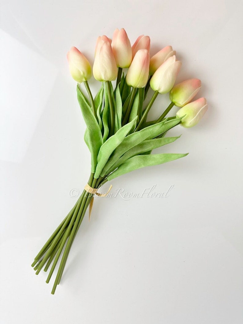 May include: A bouquet of 10 artificial tulips in a pale pink and cream colour with green stems. The stems are tied together with a brown ribbon.