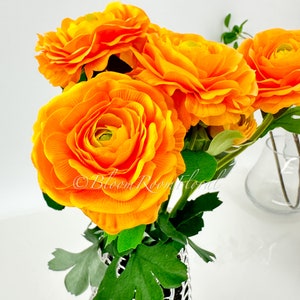 May include: A bouquet of bright orange ranunculus flowers in a silver vase with green leaves. The flowers are arranged in a cluster and are in full bloom.