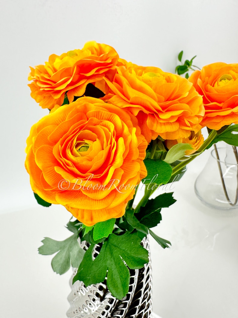 May include: A bouquet of bright orange ranunculus flowers in a silver vase with green leaves. The flowers are arranged in a cluster and are in full bloom.