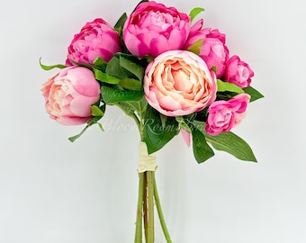 May include: A bouquet of artificial peonies in shades of pink and fuchsia, with green leaves. The flowers are tightly clustered, and the stems are bound with a beige ribbon. The arrangement is set against a white background.