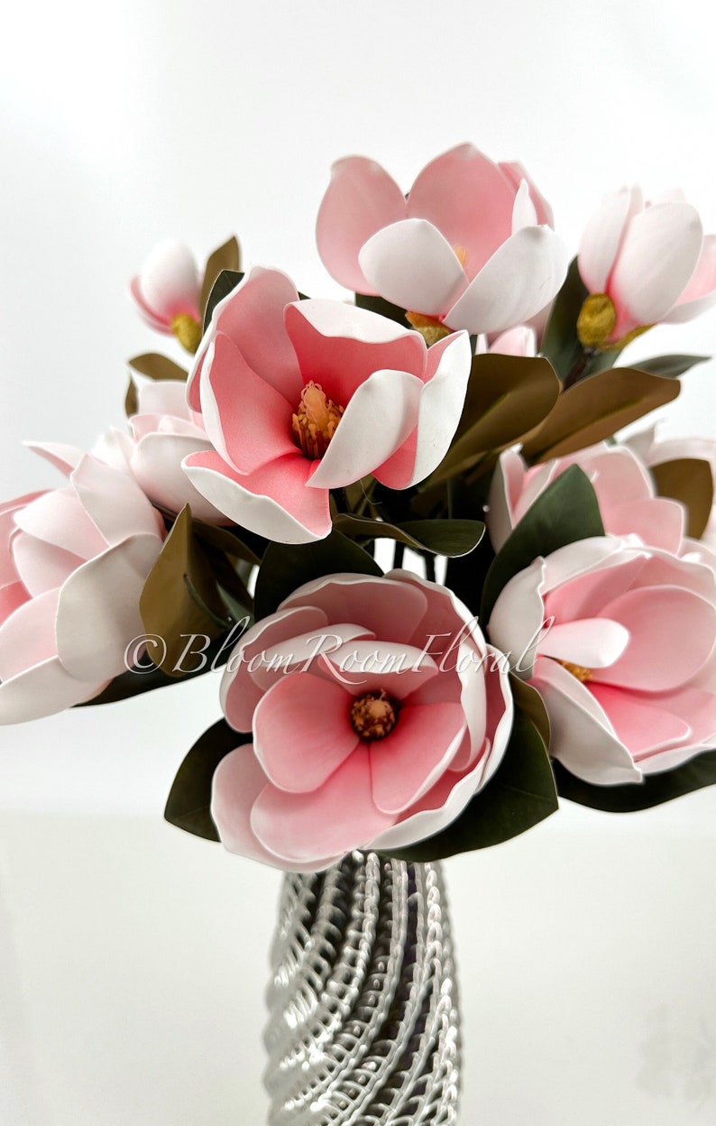 May include: A bouquet of pink and white magnolia flowers in a silver vase. The flowers are arranged in a cluster, with the stems extending out from the vase. The flowers are in full bloom, with their petals open wide. The vase is a simple, elegant design, with a silver finish.