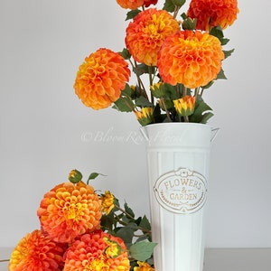 May include: A white metal vase with a gold inscription that reads "Flowers & Garden". The vase is filled with orange and yellow dahlia flowers.