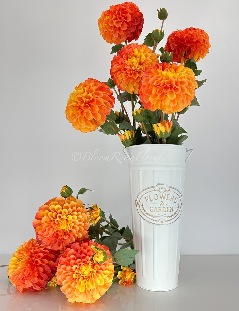 May include: A white metal vase with a gold inscription that reads "Flowers & Garden". The vase is filled with orange and yellow dahlia flowers.
