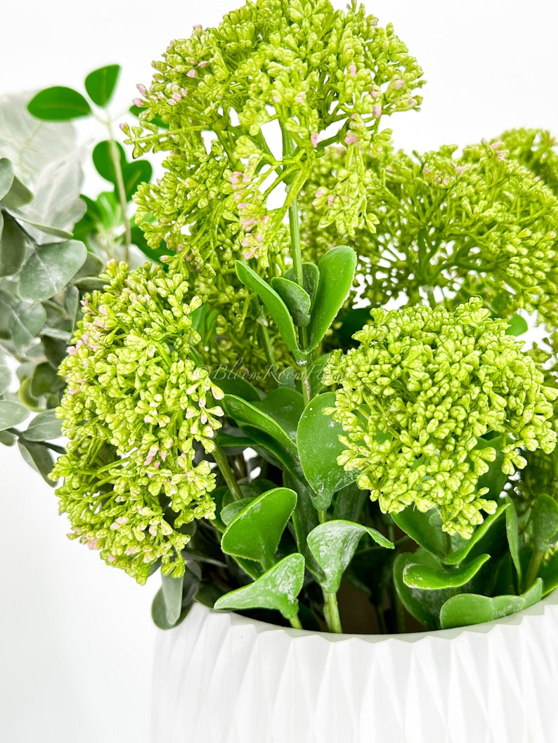 May include: A white ceramic vase with a textured surface, filled with a green and pink artificial flower arrangement.