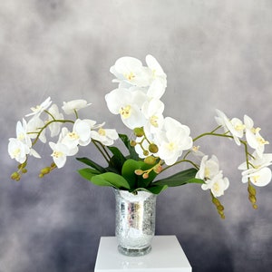 May include: A bouquet of white orchids arranged in a silver vase. The orchids are in full bloom and have delicate petals. The vase is a classic design with a textured surface.
