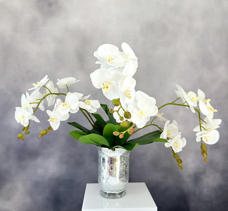 May include: A bouquet of white orchids arranged in a silver vase. The orchids are in full bloom and have delicate petals. The vase is a classic design with a textured surface.