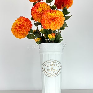 May include: A white metal vase with the words "Flowers & Garden" printed on it. The vase is filled with a bouquet of orange and yellow dahlias.