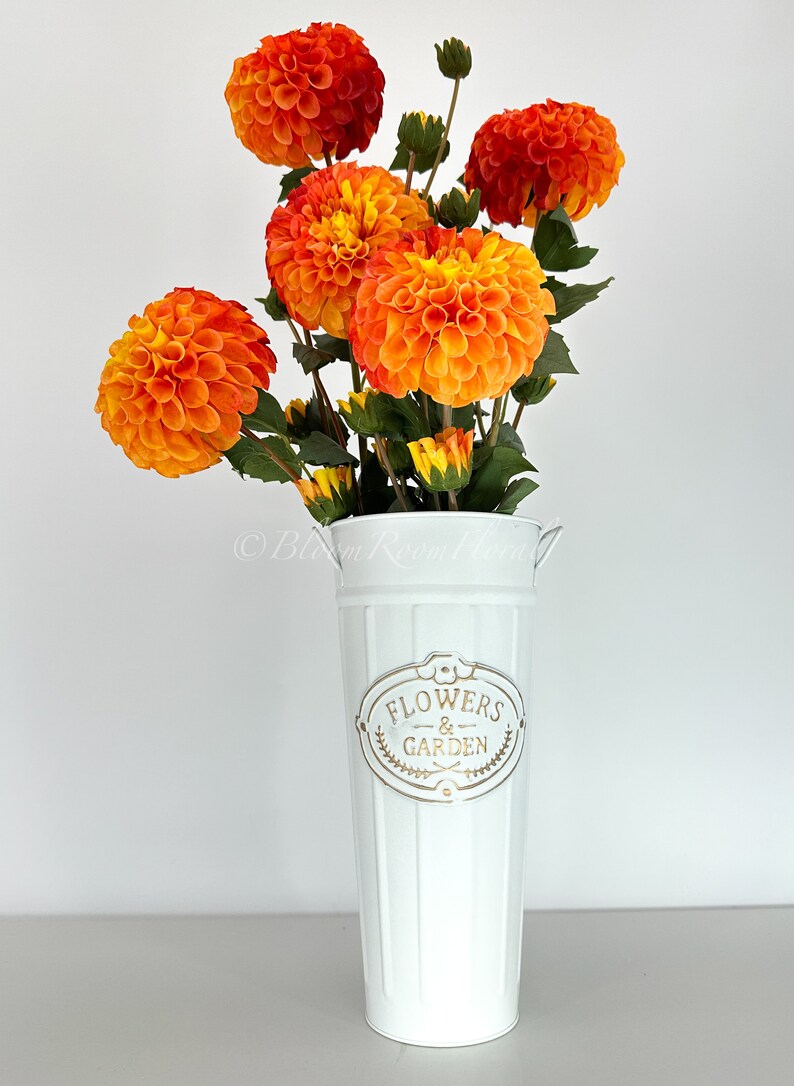 May include: A white metal vase with the words "Flowers & Garden" printed on it. The vase is filled with a bouquet of orange and yellow dahlias.