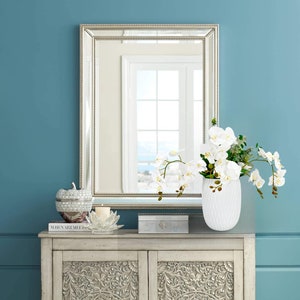 May include: A white ceramic vase with a textured surface holds a bouquet of white orchids with green leaves. The vase is on a light wood cabinet with intricate carvings. A silver framed mirror hangs on a blue wall behind the vase.