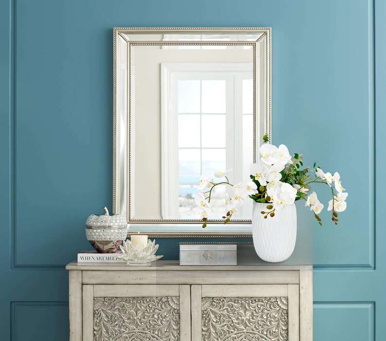 May include: A white ceramic vase with a textured surface holds a bouquet of white orchids with green leaves. The vase is on a light wood cabinet with intricate carvings. A silver framed mirror hangs on a blue wall behind the vase.