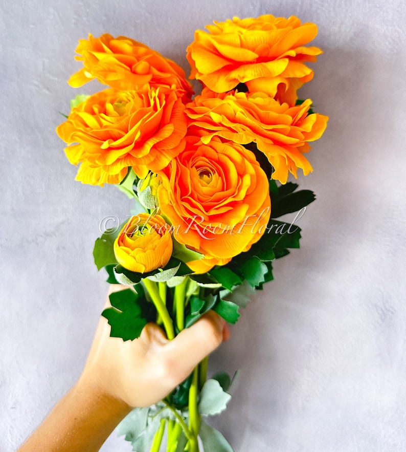 May include: A bouquet of bright orange ranunculus flowers with green stems and leaves. The flowers are arranged in a hand holding the bouquet.