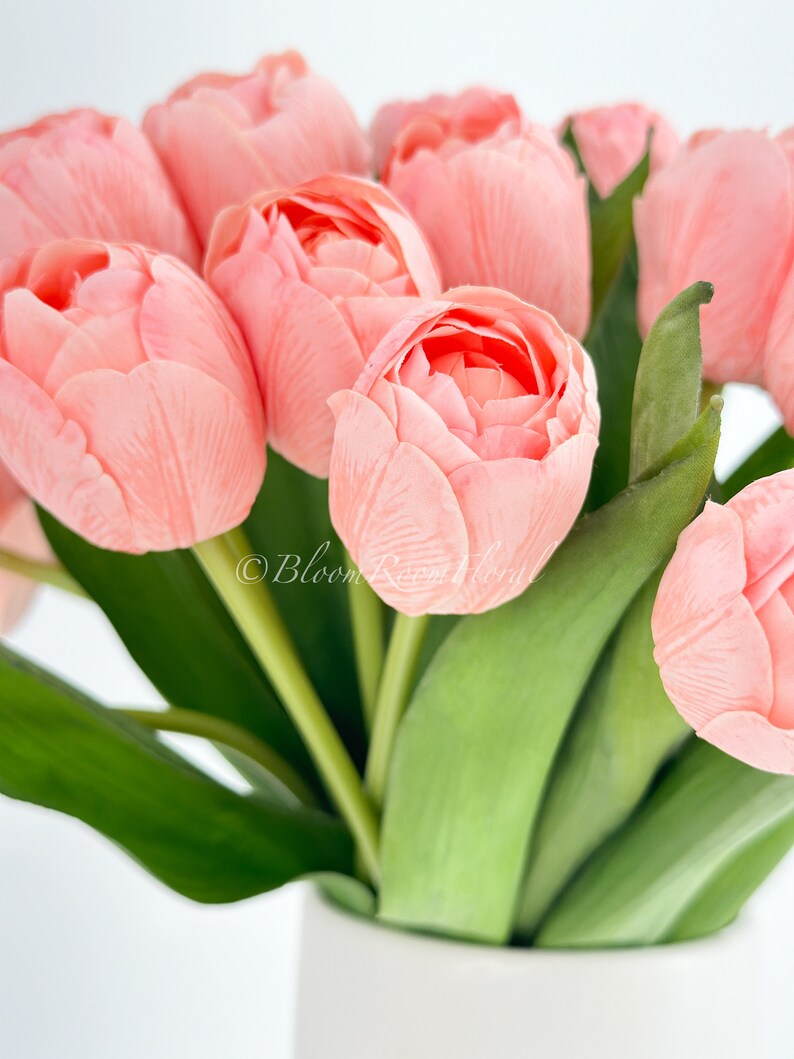 May include: A bouquet of pink tulips with green stems in a white vase. The tulips are arranged in a cluster, with the stems extending out from the center. The vase is simple and elegant, with a smooth white finish.