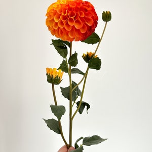 May include: A single stem of orange and yellow dahlia flowers with green leaves. The flower is in bloom and has a round shape.