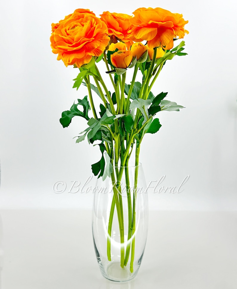 May include: A bouquet of orange ranunculus flowers in a clear glass vase. The flowers are arranged in a simple, elegant style.