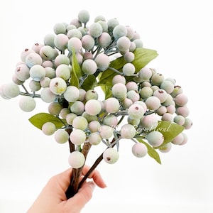 以下が含まれることがあります： 薄い緑色とピンク色のベリーが付いた、フェイクベリーの茎の束。茎は茶色で、緑の葉が付いています。