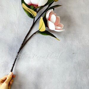 May include: A bouquet of three pink magnolia flowers with green leaves and brown stems. The flowers are arranged in a vase and the stems are visible. The background is a light gray.
