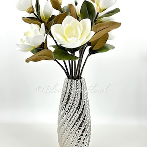 K&ouml;nnte beinhalten: Eine silberne Vase mit einem strukturierten, wirbelnden Design h&auml;lt einen Strau&szlig; k&uuml;nstlicher Magnolienbl&uuml;ten. Die Bl&uuml;ten sind wei&szlig; mit einem gelben Zentrum, und die Bl&auml;tter sind gr&uuml;n und braun. Die Vase steht auf einer wei&szlig;en Oberfl&auml;che.