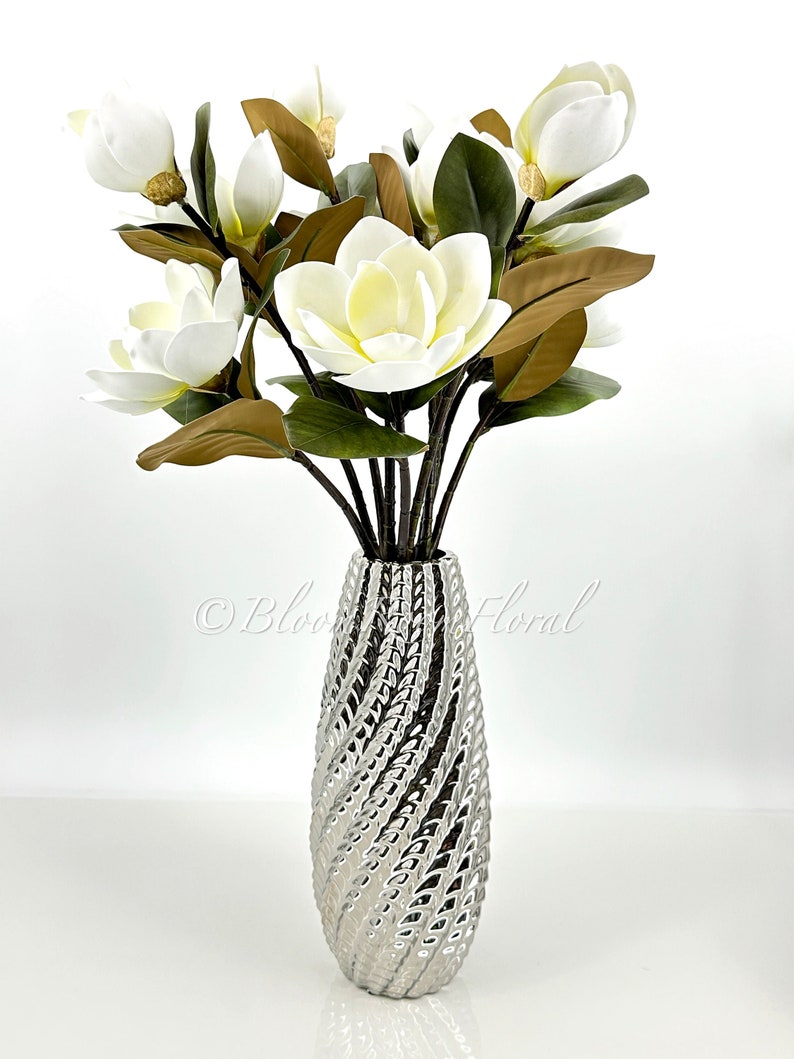 K&ouml;nnte beinhalten: Eine silberne Vase mit einem strukturierten, wirbelnden Design h&auml;lt einen Strau&szlig; k&uuml;nstlicher Magnolienbl&uuml;ten. Die Bl&uuml;ten sind wei&szlig; mit einem gelben Zentrum, und die Bl&auml;tter sind gr&uuml;n und braun. Die Vase steht auf einer wei&szlig;en Oberfl&auml;che.