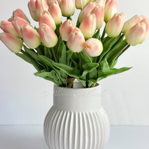 May include: A bouquet of pink and white artificial tulips in a white ceramic vase with vertical ridges. The vase has the text "Bloom Room Floral" on the side.