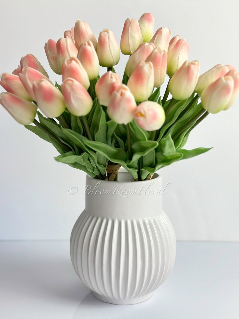 May include: A bouquet of pink and white artificial tulips in a white ceramic vase with vertical ridges. The vase has the text "Bloom Room Floral" on the side.