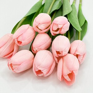 Puede incluir: Un ramo de tulipanes artificiales rosas. Los p&eacute;talos muestran vetas sutiles. Los tallos y hojas verdes contrastan con el fondo blanco. Estas flores artificiales ofrecen un toque primaveral, ideal para decoraci&oacute;n.