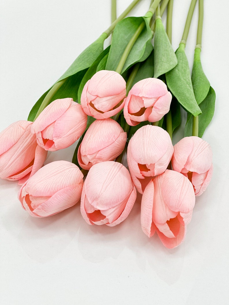 Puede incluir: Un ramo de tulipanes artificiales rosas. Los p&eacute;talos muestran vetas sutiles. Los tallos y hojas verdes contrastan con el fondo blanco. Estas flores artificiales ofrecen un toque primaveral, ideal para decoraci&oacute;n.