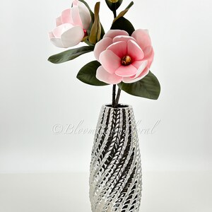 May include: A silver ceramic vase with a textured swirl design, holding a bouquet of three pink magnolia flowers.