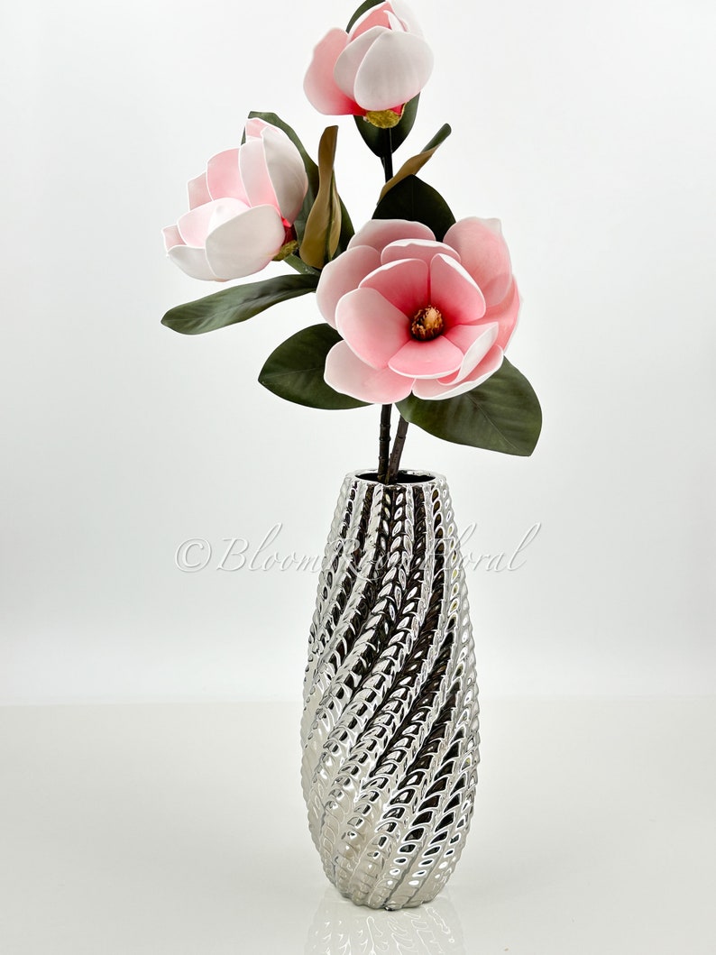 May include: A silver ceramic vase with a textured swirl design, holding a bouquet of three pink magnolia flowers.