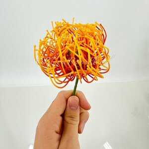 K&ouml;nnte beinhalten: Eine einzelne k&uuml;nstliche Blume in Orange- und Gelbt&ouml;nen mit einem langen Stiel. Die Blume besteht aus d&uuml;nnen, gekr&auml;uselten Dr&auml;hten, die eine fluffige, kugelf&ouml;rmige Form bilden.