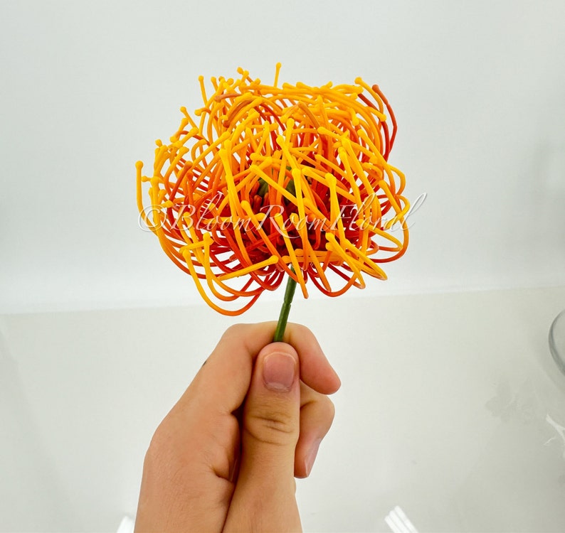 K&ouml;nnte beinhalten: Eine einzelne k&uuml;nstliche Blume in Orange- und Gelbt&ouml;nen mit einem langen Stiel. Die Blume besteht aus d&uuml;nnen, gekr&auml;uselten Dr&auml;hten, die eine fluffige, kugelf&ouml;rmige Form bilden.