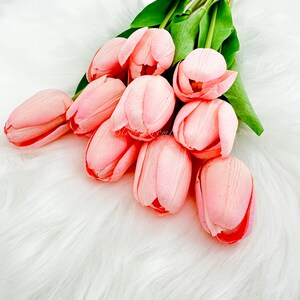 Puede incluir: Un ramo de tulipanes rosas artificiales con tallos y hojas verdes. Los tulipanes est&aacute;n dispuestos sobre una superficie blanca y esponjosa. Los p&eacute;talos son de color rosa suave y las flores est&aacute;n en varias etapas de floraci&oacute;n.