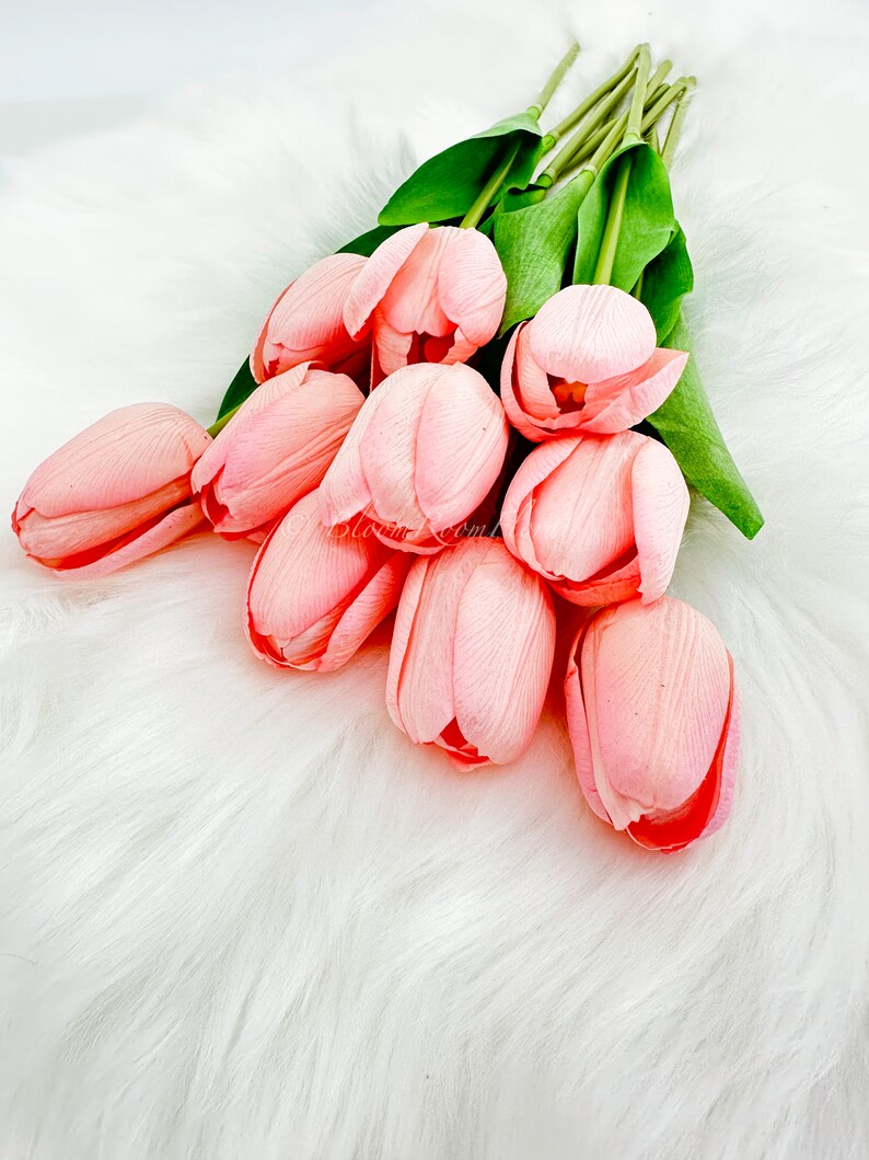 Puede incluir: Un ramo de tulipanes rosas artificiales con tallos y hojas verdes. Los tulipanes est&aacute;n dispuestos sobre una superficie blanca y esponjosa. Los p&eacute;talos son de color rosa suave y las flores est&aacute;n en varias etapas de floraci&oacute;n.