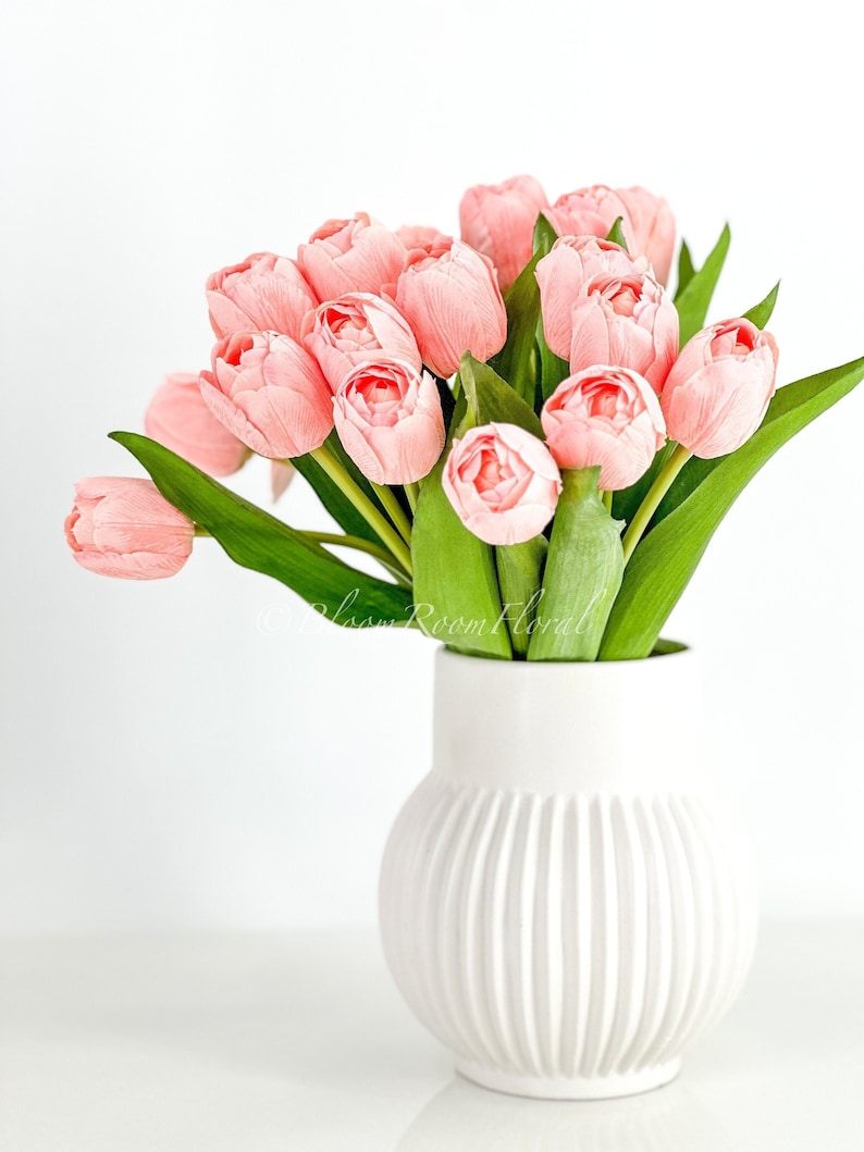 May include: A bouquet of pink tulips in a white ceramic vase with vertical ridges. The vase is on a white surface.