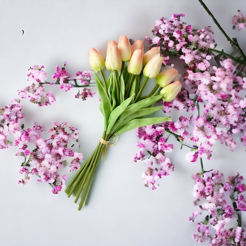 May include: A bouquet of peach-coloured artificial tulips with green stems and leaves. The tulips are arranged in a fan shape and are surrounded by pink flowers.