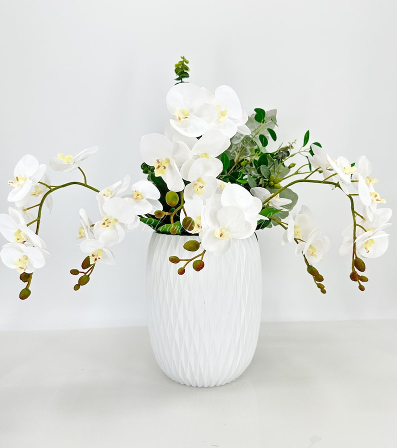 May include: White orchid flower arrangement in a white textured vase. The orchids are arranged in a cascading style with green leaves.
