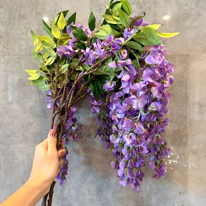 May include: A bunch of artificial wisteria flowers with purple blooms and green leaves. The stems are brown and the flowers are hanging down.
