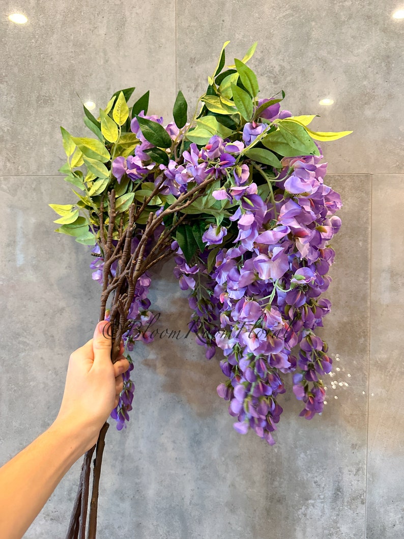 May include: A bunch of artificial wisteria flowers with purple blooms and green leaves. The stems are brown and the flowers are hanging down.
