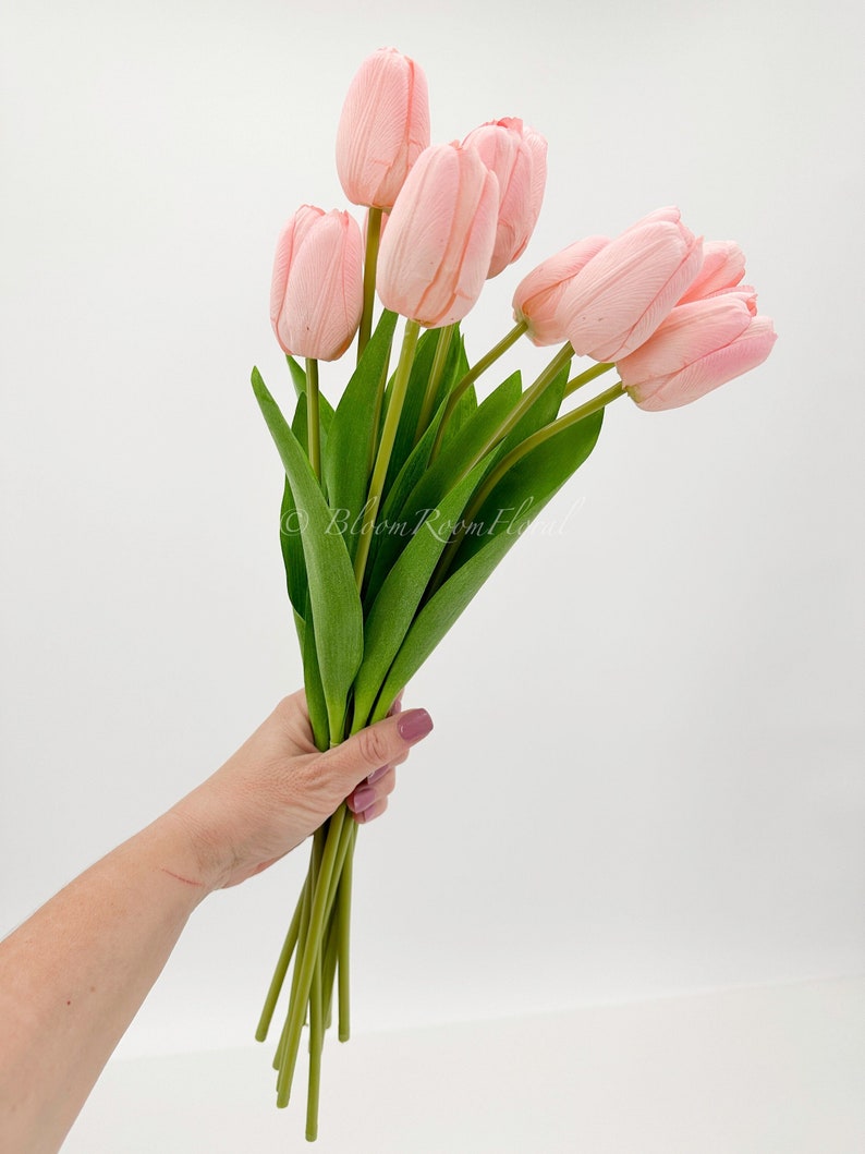 Puede incluir: Un ramo de tulipanes rosas artificiales con tallos y hojas verdes. Los tulipanes se sostienen en una mano sobre un fondo blanco. La imagen incluye el texto "Bloom Room Floral".