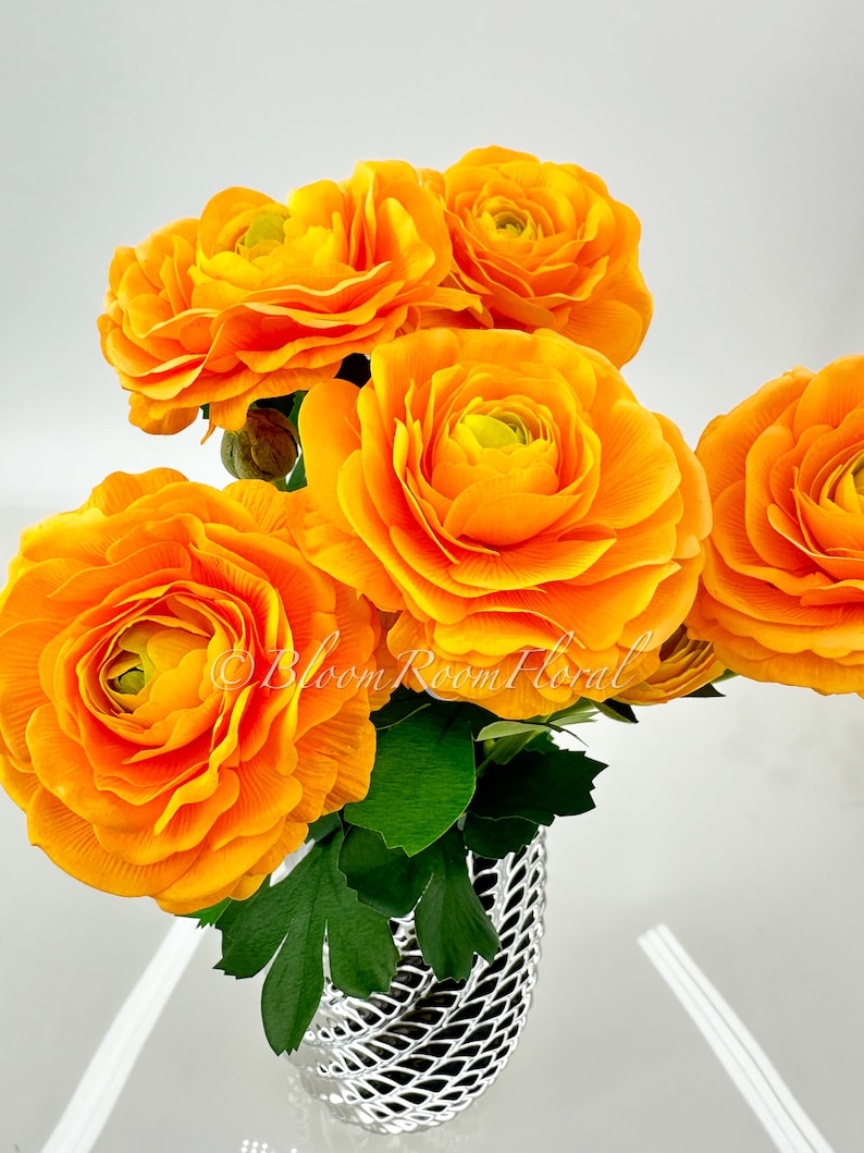 May include: A bouquet of bright orange ranunculus flowers in a silver vase. The flowers are arranged in a loose, natural style. The vase has a textured, woven design.