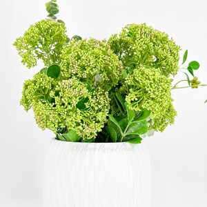 May include: A white ceramic vase with a textured surface filled with a bouquet of green artificial flowers.