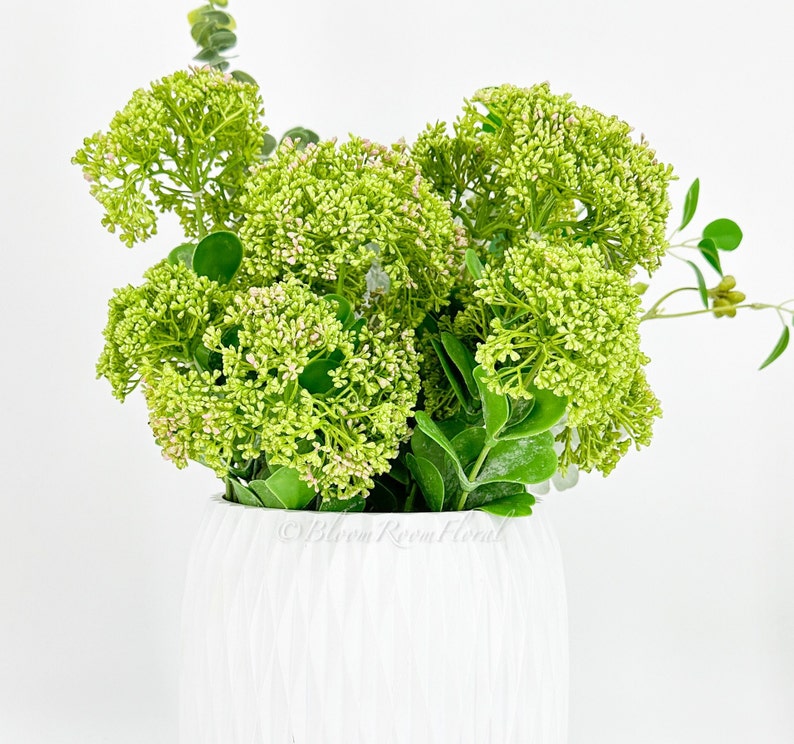 May include: A white ceramic vase with a textured surface filled with a bouquet of green artificial flowers.