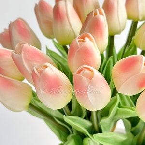 May include: A bouquet of peach-coloured artificial tulips with green leaves in a white vase. The tulips are arranged in a cluster, with some of the flowers facing the camera and others facing away. The leaves are arranged around the tulips, creating a lush and full bouquet.